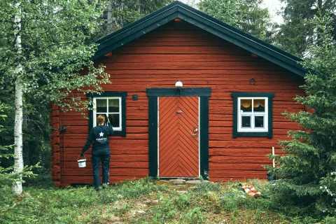 Person painting window frame of a red wooden cabin surrounded by green forest during daytime.