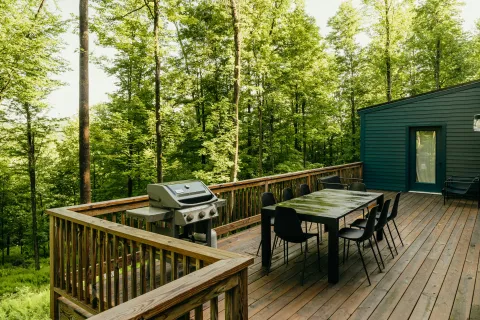 Wooden deck with dining table, chairs, and grill surrounded by dense green forest on a sunny day