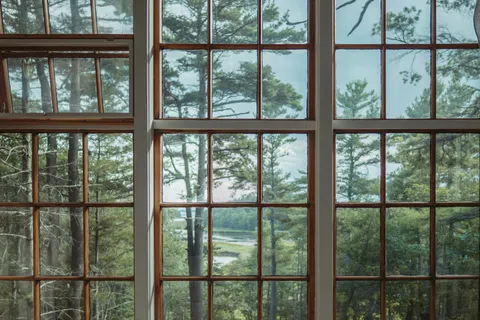 View of tall pine trees and a river seen through large wooden framed window panes with curtains.
