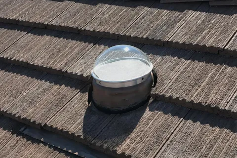 Clear dome skylight installed on textured brown roof tiles casting shadows in sunlight