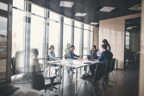 Business team in formal attire holding a meeting around a large table in a bright office with glass walls.