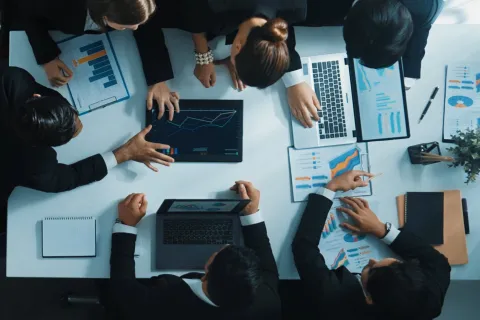 Business team analyzing graphs and charts on laptops and printed reports during a meeting at a white table.