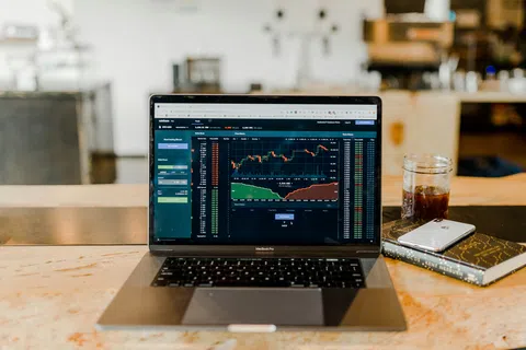 Laptop on wooden table displaying stock market charts with a phone, book, and glass nearby in a cozy setting