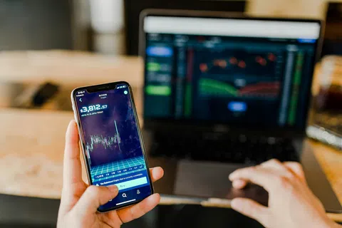 Person holding smartphone with Bitcoin price chart while using laptop in the background on wooden table