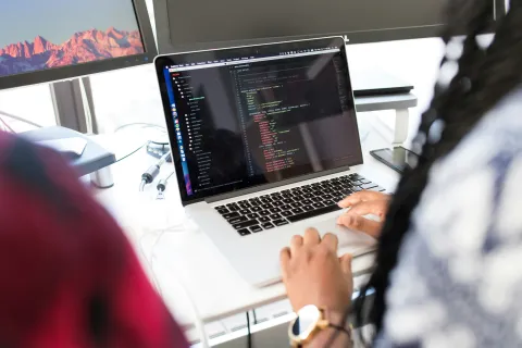 Person coding on a laptop with visible code editor and a monitor showing a mountain wallpaper in a modern office.