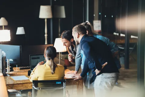 Team of four professionals collaborating and reviewing work on a laptop in a modern office setting with warm lighting.