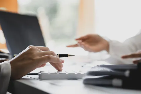 Hands of two professionals working with calculator and documents at office desk near laptop.