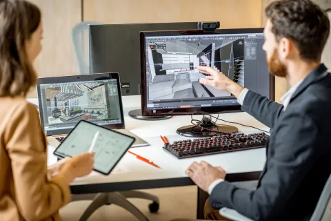 Two architects collaborate on 3D interior design using computer and tablet at office desk