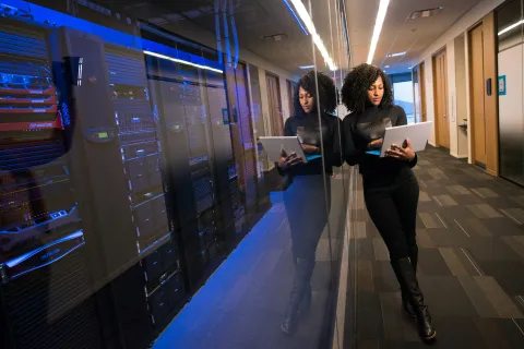 Woman with curly hair using laptop in a server room with reflections on glass wall and tech equipment