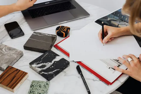 Designer sketching building plans surrounded by marble and wood samples on a white table.