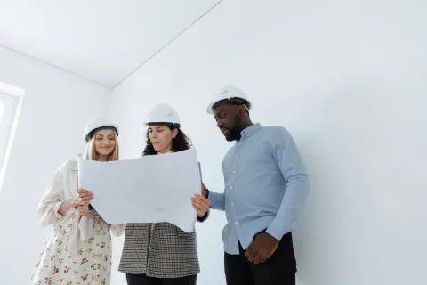 Three architects wearing helmets reviewing building blueprints in a bright, empty room with white walls.