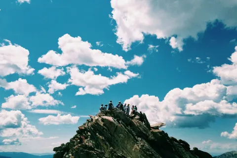 Group of hikers standing atop a rocky mountain peak against a bright blue sky with fluffy clouds.