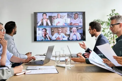 A diverse group in a meeting, engaging in a video conference displayed on a screen.