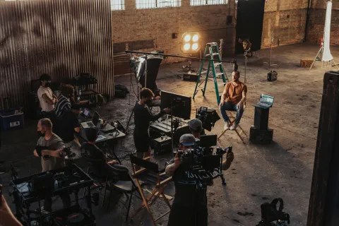 Film crew working on set with director, camera operators, lighting, and actor in a spacious industrial studio.