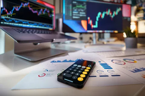 Smartphone with calculator app on desk surrounded by financial charts and stock market graphs on screens.