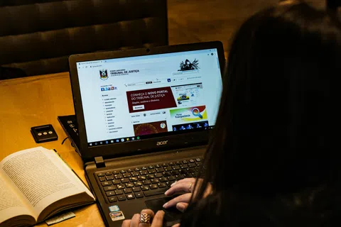 Person browsing the Tribunal de Justiça website on an Acer laptop at a wooden table with an open book.