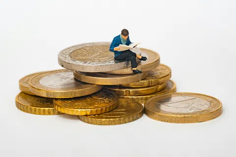 Miniature figure reading a book while sitting on a stack of Euro coins representing finance and knowledge.