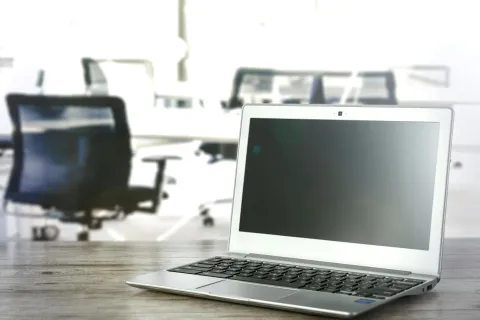 Silver laptop with black keyboard on wooden desk in bright modern office with blurred chairs in background