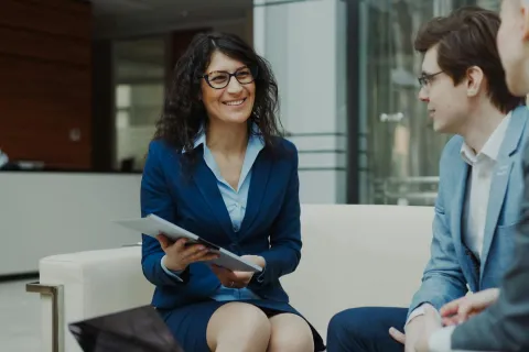 Smiling businesswoman holding documents while discussing with two male colleagues in modern office lounge.