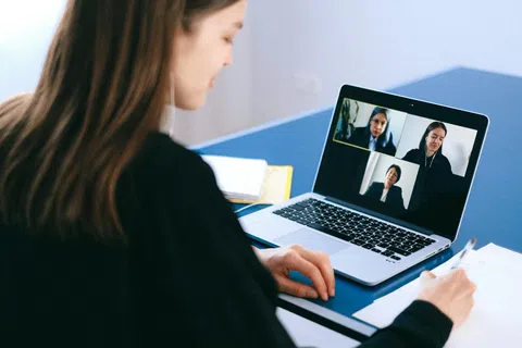 Woman attending virtual meeting on laptop while taking notes at blue desk in bright room