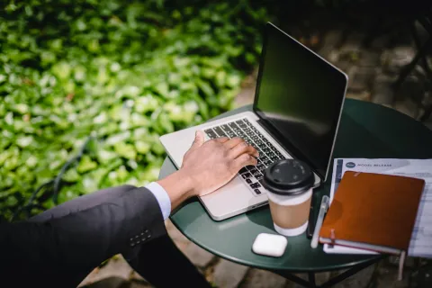 Person in suit working on laptop outdoors with coffee, notebook, AirPods, and documents on table.