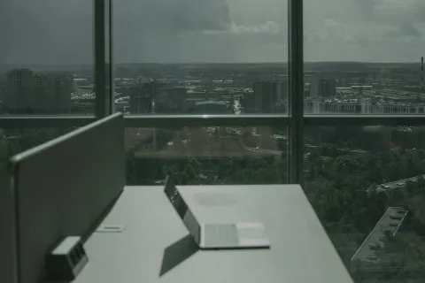 Empty office desk with laptop overlooking a cityscape through large glass windows on a cloudy day