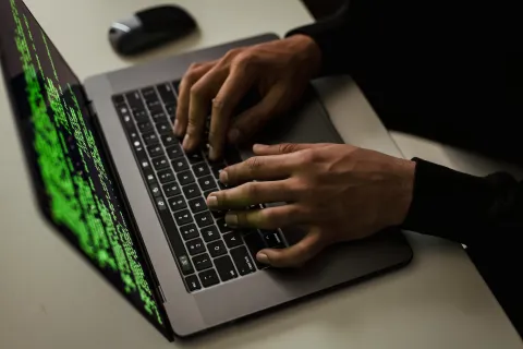 Hands typing on a laptop keyboard with green code on the screen and a wireless mouse nearby.