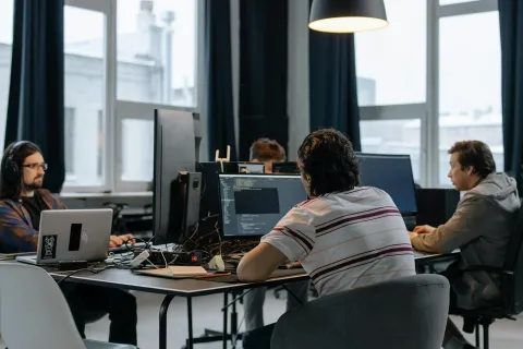 Four software developers working on computers in a modern office with large windows and black curtains