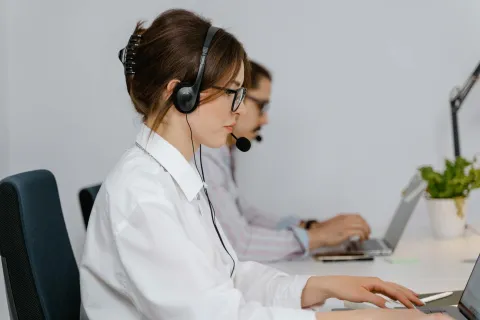 Two call center agents wearing headsets working on laptops in a modern office setting.