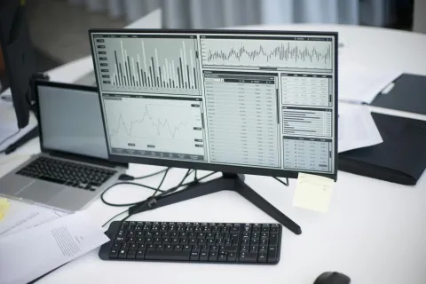 Computer monitor on desk displaying charts, graphs, and data with keyboard and laptop nearby in office setting.