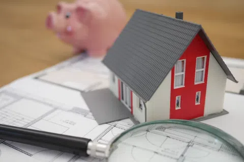 Miniature house model on architectural plans with magnifying glass and pink piggy bank in the background