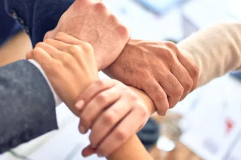 Four diverse hands grasp each other's wrists forming a unity symbol in a business setting.