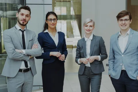 Four diverse business professionals in formal attire standing confidently in a modern office lobby.