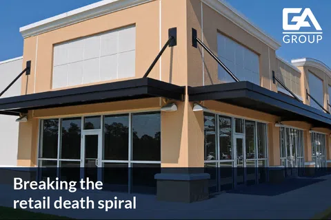 Empty modern retail strip mall storefronts with large glass windows and beige exterior under clear blue sky.