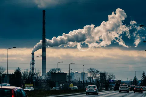 Smoke billowing from factory chimneys against a cloudy sky over a busy roadside with cars.