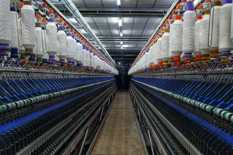 Rows of industrial textile machines with white thread spools in a spacious, well-lit factory corridor.