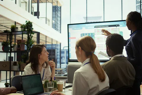 Team meeting in modern office with diverse professionals discussing data on large screen display.