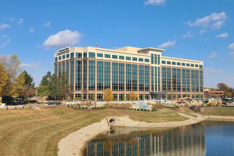 Modern office building with large glass windows by a pond under a blue sky with clouds and autumn trees nearby