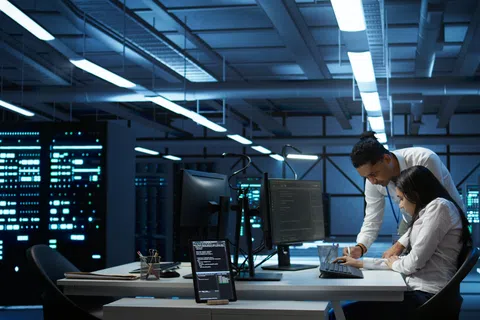 Two IT professionals work together on coding at a desk in a dimly lit modern data center with server racks.