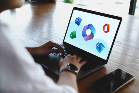 Person using laptop displaying Microsoft Office app icons on screen with smartphone nearby on wooden table