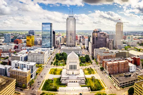 Aerial view of a city skyline with prominent skyscrapers and a central historic monument surrounded by green lawns under a blue sky.