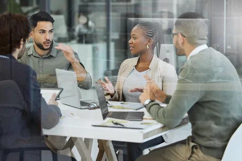 Diverse team in business casual discussing ideas around a table with laptops and documents in a modern office.