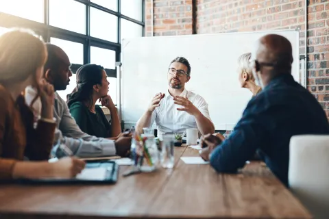 Diverse group of professionals engaged in a meeting with speaker explaining ideas around a table in modern office.