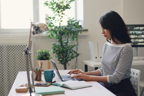 Young woman working on a laptop at a tidy desk with plants, books, and a mug in a bright home office.