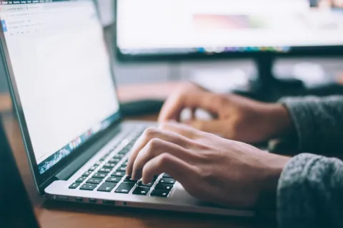 Person typing on laptop keyboard with computer screens in the background in a workspace setting