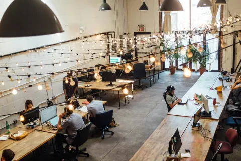Modern coworking space with people working at wooden desks under hanging string lights and large windows.
