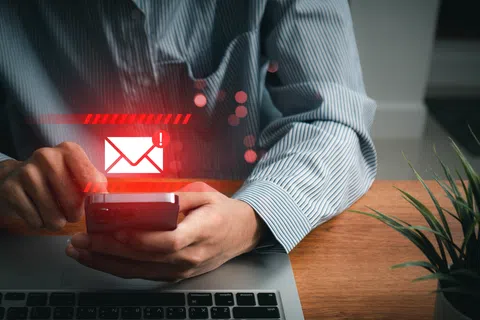 Person holding a smartphone with a red email notification icon above, sitting at a laptop on a wooden desk.