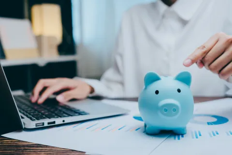 Person working on laptop with blue piggy bank and financial charts on desk symbolizing savings and finance.