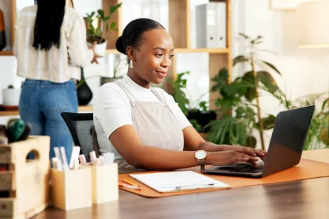 Woman in apron working on laptop at plant-filled home office desk with organized supplies