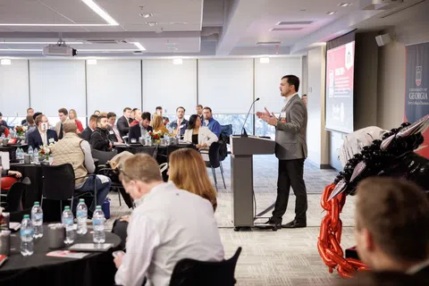 Speaker presenting at a business conference with attendees seated at round tables in a modern venue.